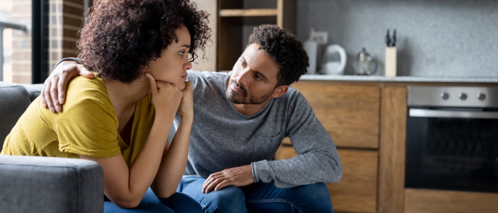 women and man sitting on a sofa, he puts his arm around her 