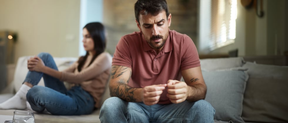 Man is sitting with his back to a woman, looking insecurely at his fingers. 