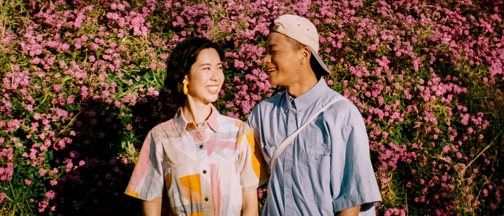 A woman and a man standing next to each other in front of flowers. They smile at each other and seem to be attracted to each other.