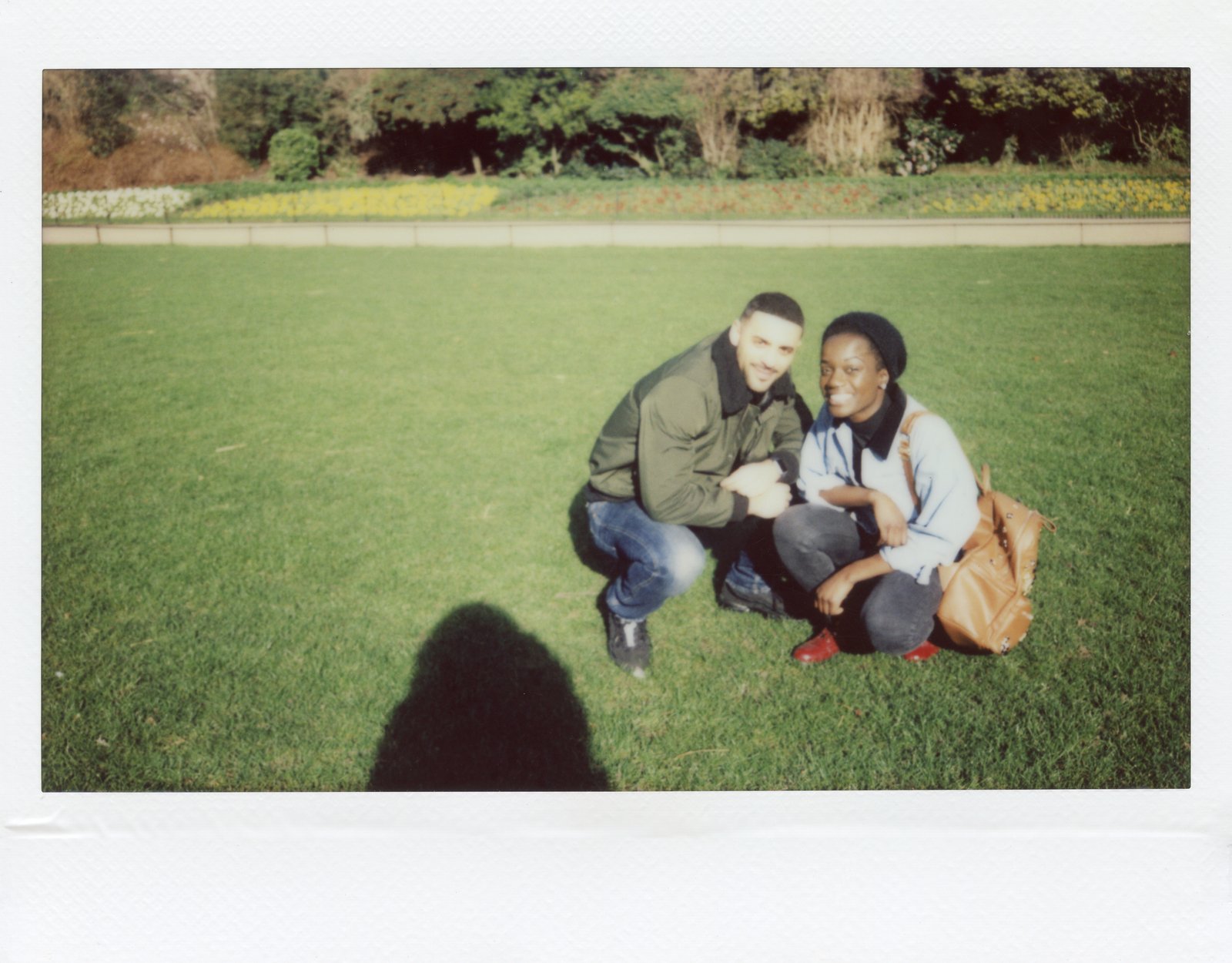 A polaroid-style grainy photo with a man and woman wearing denim pants and jackets sitting in a grassy park smiling at the camera. 