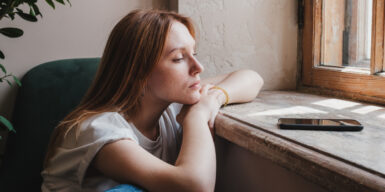 A woman experiencing unrequited love, looking at her phone while sitting at a window.